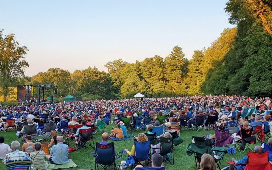 An outdoor concert overlooking a meadow with people sitting in chairs looking at the stage.